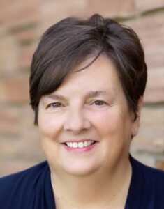 Smiling woman with short dark hair wearing a navy top, brick wall behind her (portrait).
