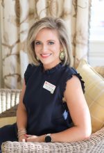 Shelley Whitehead PhD – Executive Director Smiling woman with short blonde hair wearing a navy sleeveless top and large gold earrings, seated in a wicker chair indoors.